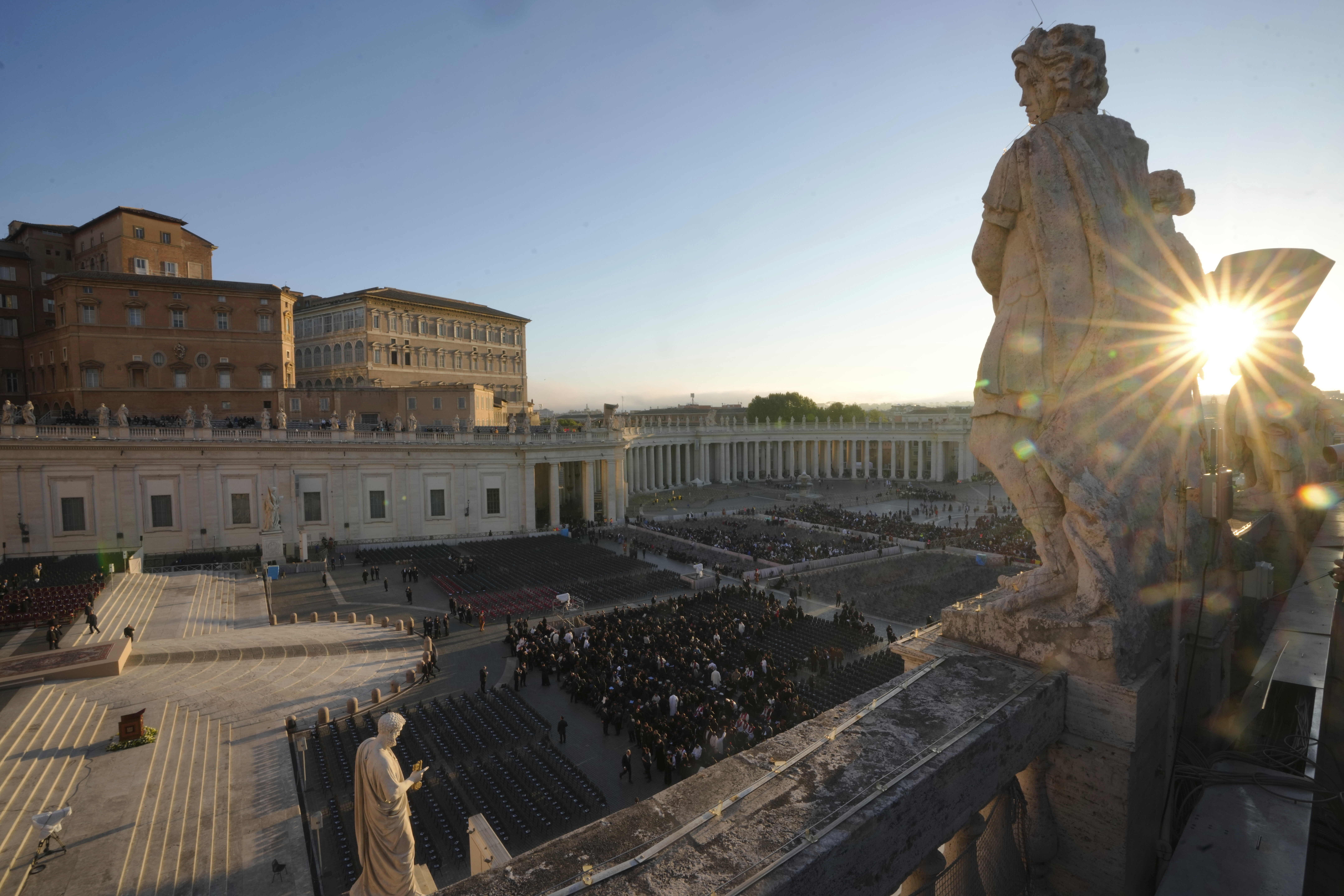 The sun rises through a statue as people begin to take their seats in St. Peter's Square, ahead of the funeral of Pope Francis at the Vatican, Saturday, April 26, 2025. (AP Photo/Markus Schreiber)